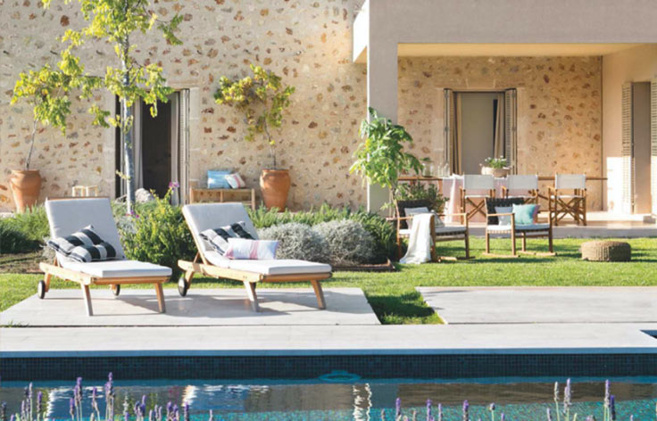 Fachada de piedra clara con tumbonas de madera, césped, piscina con flores lilas y comedor exterior cubierto