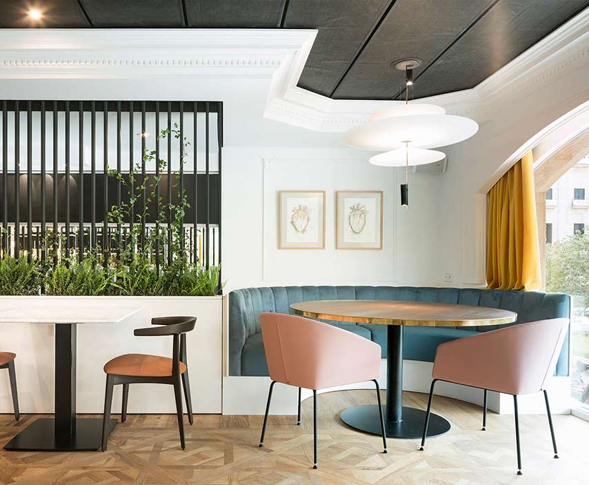 Curved blue banquette, round table and pink chairs beside a planter of ferns and an arched window
