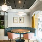 Corner with blue banquette, round table with gold edge and pink chairs, pendant lamps and yellow curtain