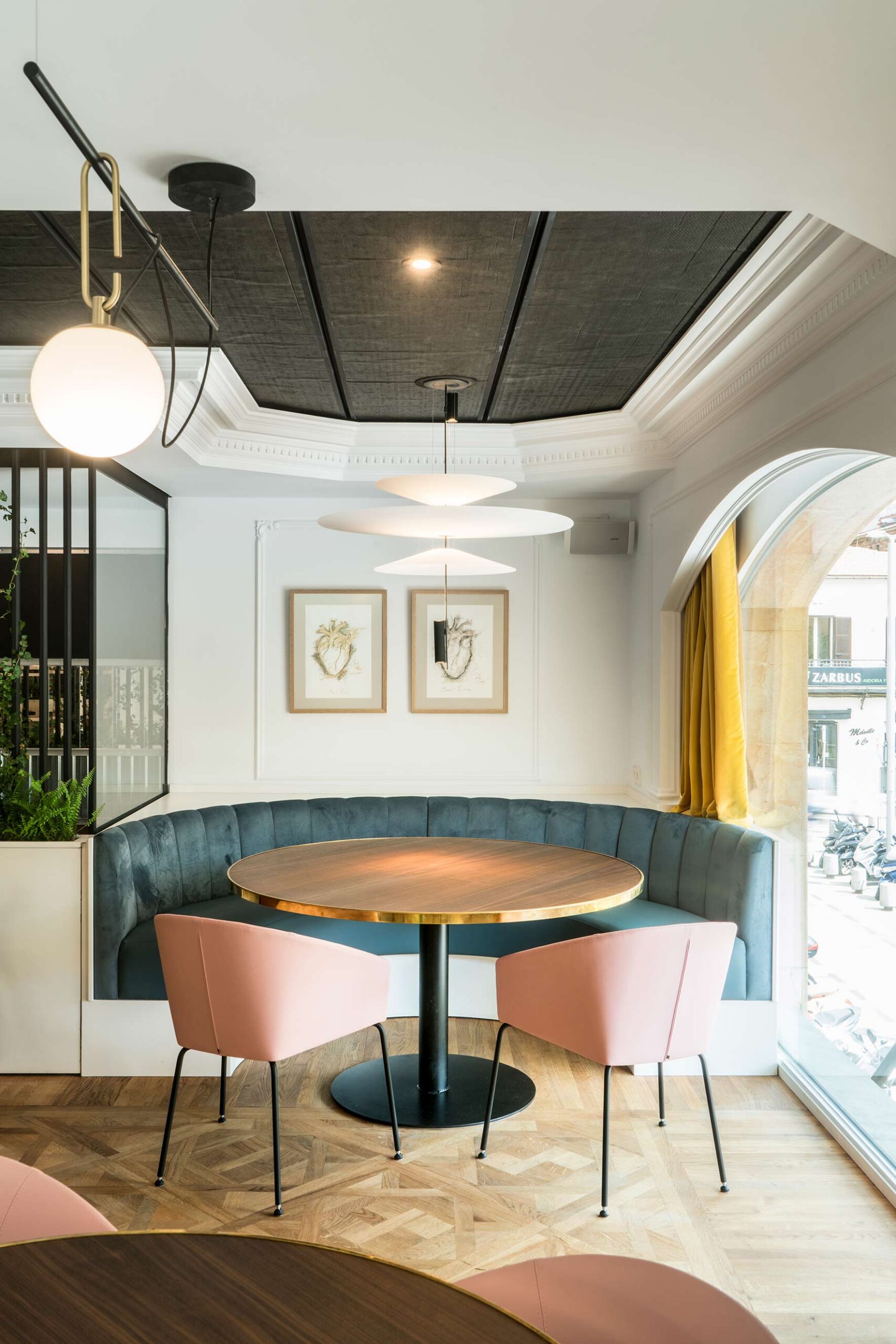 Corner with blue banquette, round table with gold edge and pink chairs, pendant lamps and yellow curtain