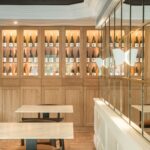Lit display cabinets in pale wood with wine bottles, beside marble tables, chairs and a mirror