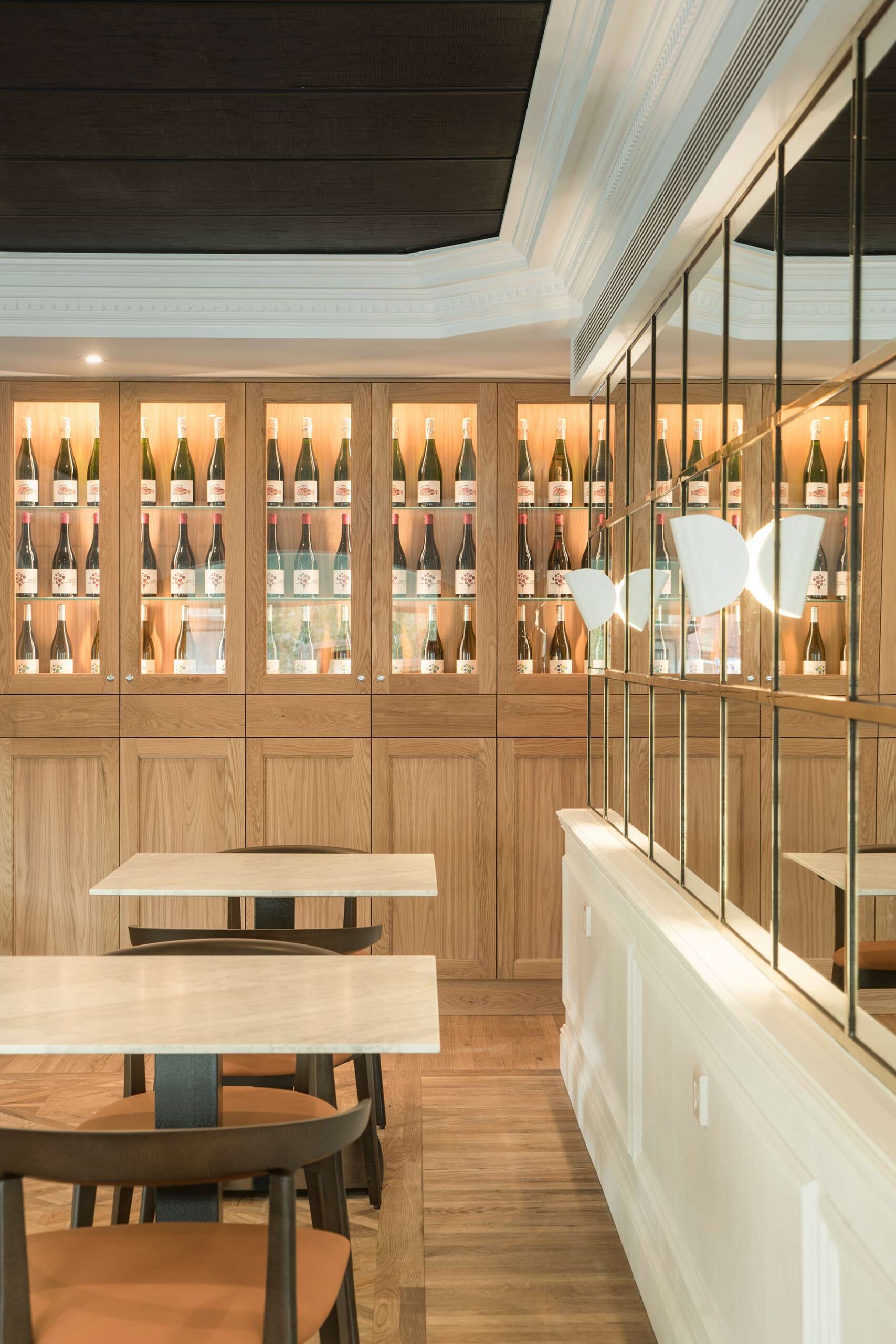 Lit display cabinets in pale wood with wine bottles, beside marble tables, chairs and a mirror