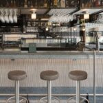 Three steel stools with brown seats in front of the bar, with bottles and hanging glasses