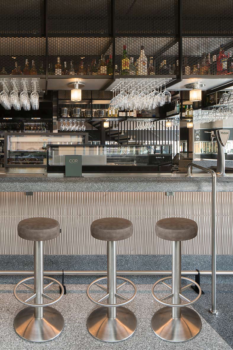 Three steel stools with brown seats in front of the bar, with bottles and hanging glasses