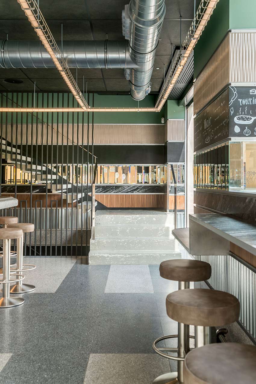 Pale stone steps between a black-barred railing and a bar with stools, beneath a linear light fitting