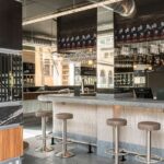 Stone bar with stools, hanging glasses, black mesh shelves and exposed ventilation ducts