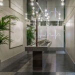 Potted palms beside the continuous washbasin, with grey chequered stone flooring and mirrors beyond