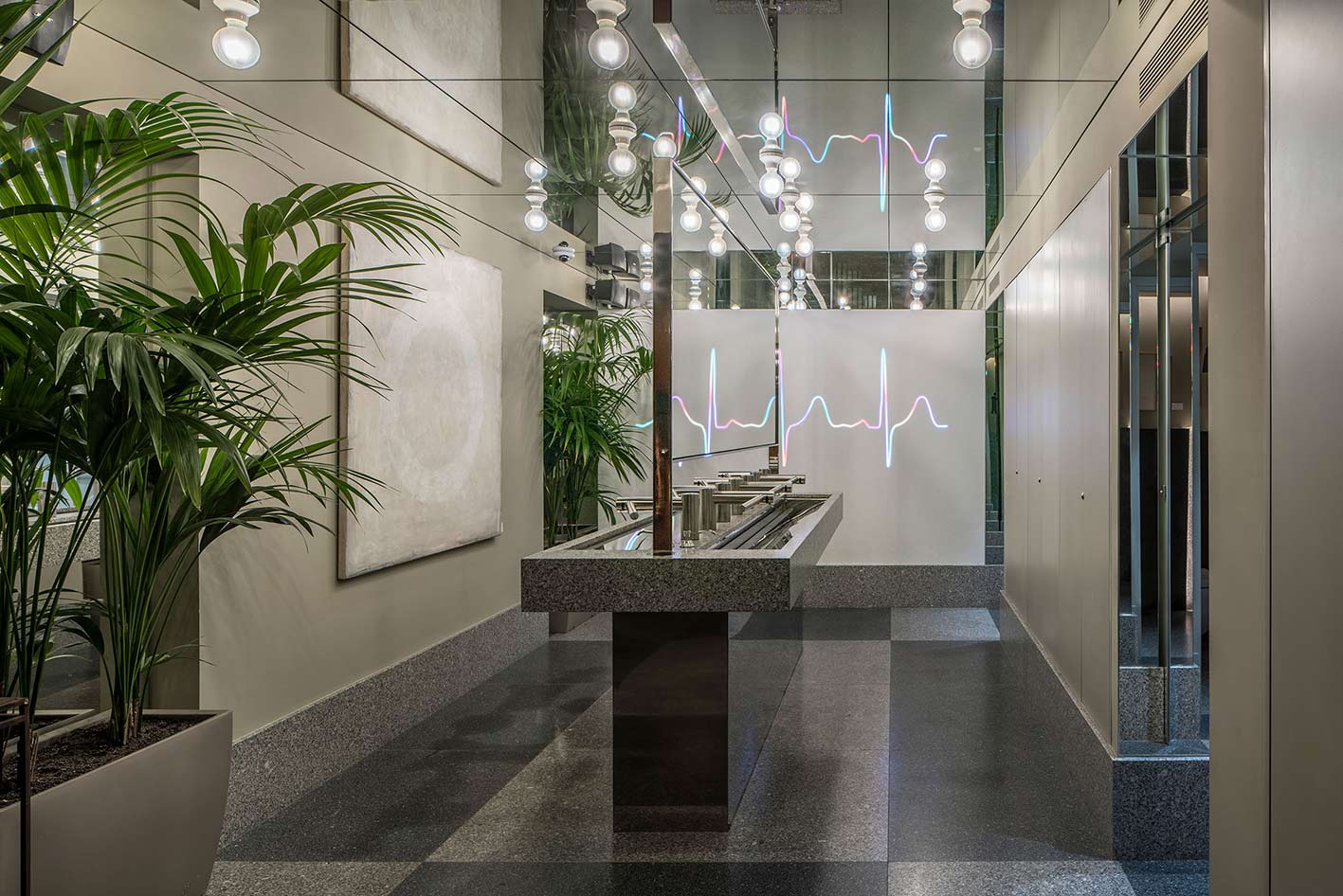 Potted palms beside the continuous washbasin, with grey chequered stone flooring and mirrors beyond