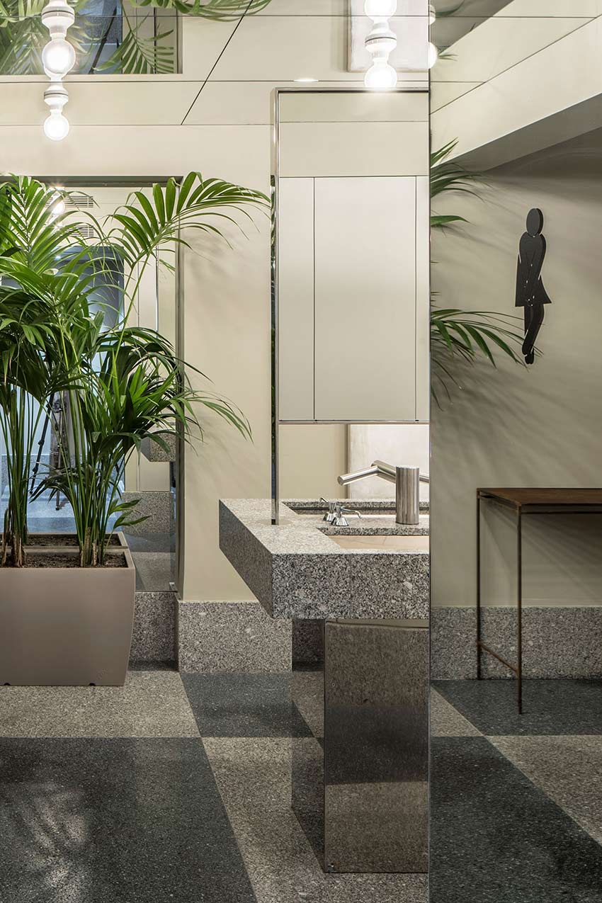 Grainy stone washbasin with mirrored pillar, potted palms and grey chequered floor