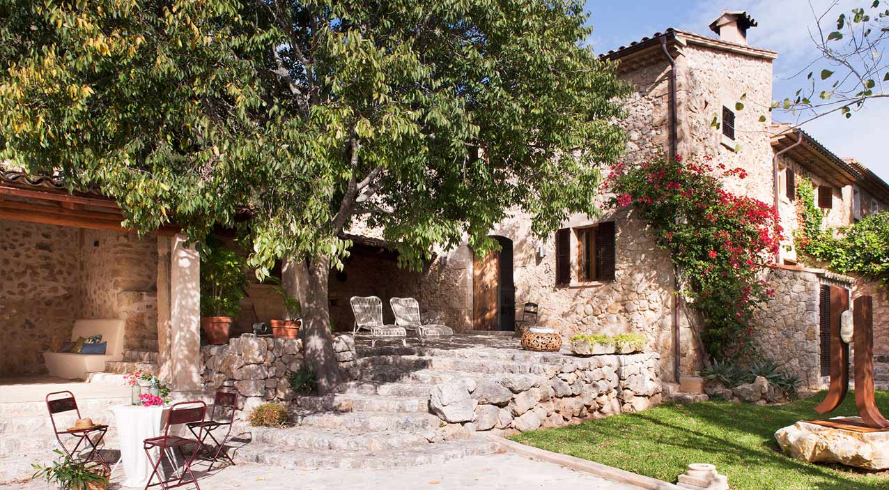 Fachada de piedra de Can Suau con trepadora en flor, escalera exterior y un gran árbol