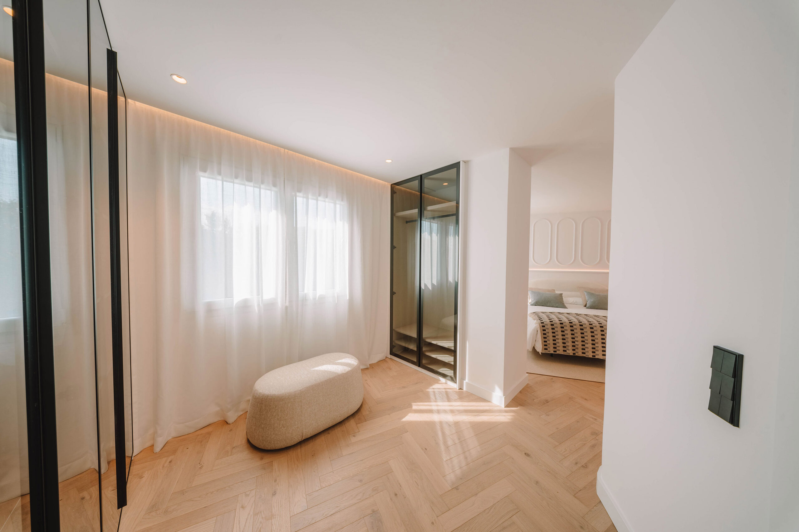 Vestidor junto al dormitorio con armario de cristal y perfilería negra, cortinas de gasa y puf beige