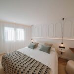 Dormitorio con cabecero de molduras ovaladas, luz indirecta, cortinas de gasa y ropa de cama blanca
