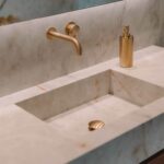 Detalle de baño con lavabo integrado en piedra clara veteada y grifería empotrada dorada