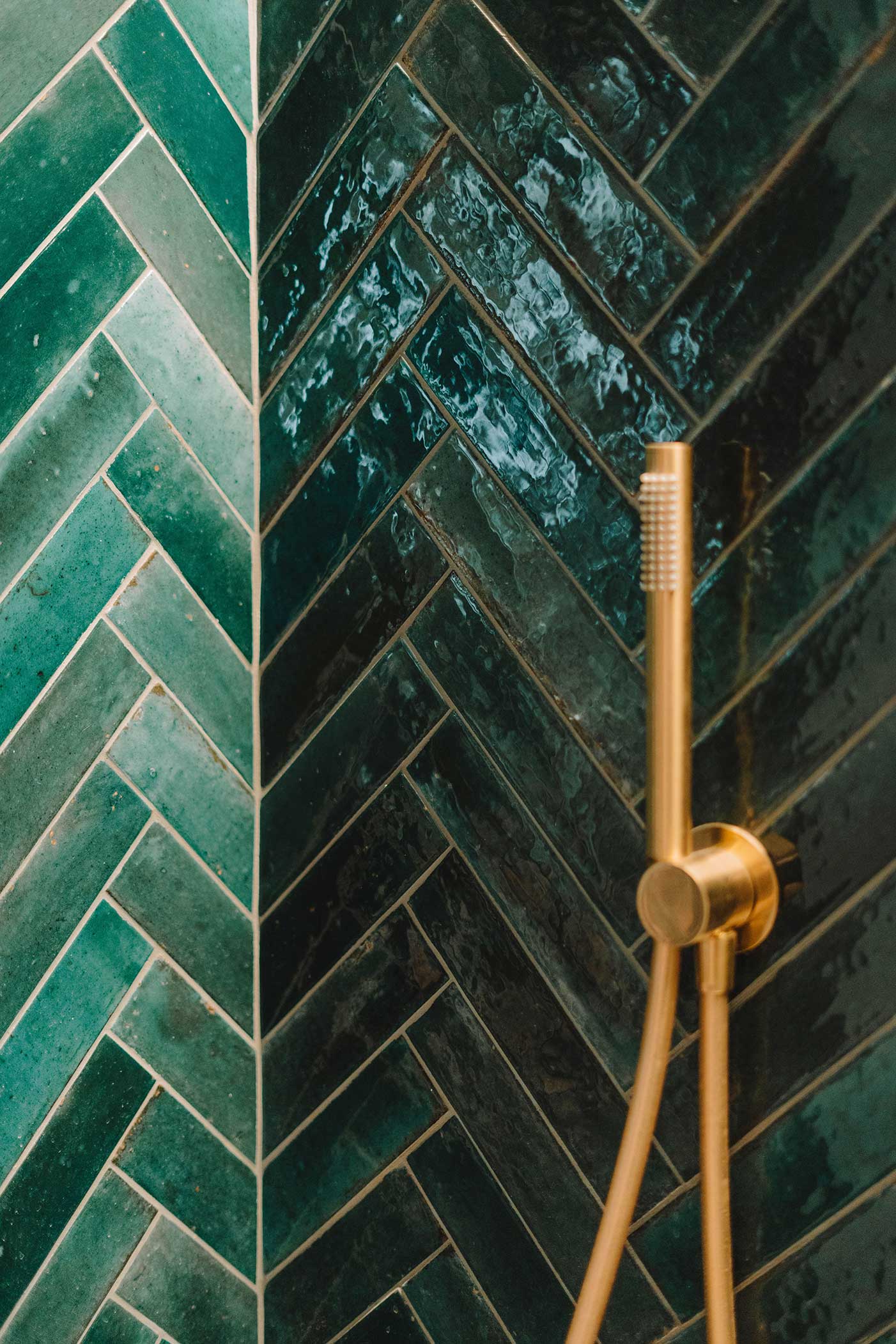 Detalle de pared de ducha con azulejos verdes esmaltados en espiga y ducha de mano dorada
