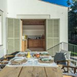 Terrasse mit gedecktem Holztisch, olivgrünen Fensterläden zur Küche hin geöffnet und Rasen im Hintergrund