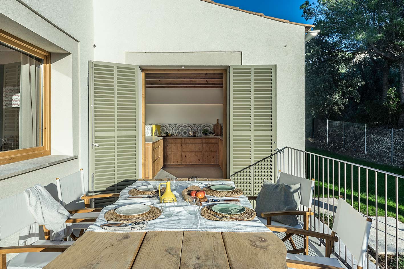 Terrasse mit gedecktem Holztisch, olivgrünen Fensterläden zur Küche hin geöffnet und Rasen im Hintergrund