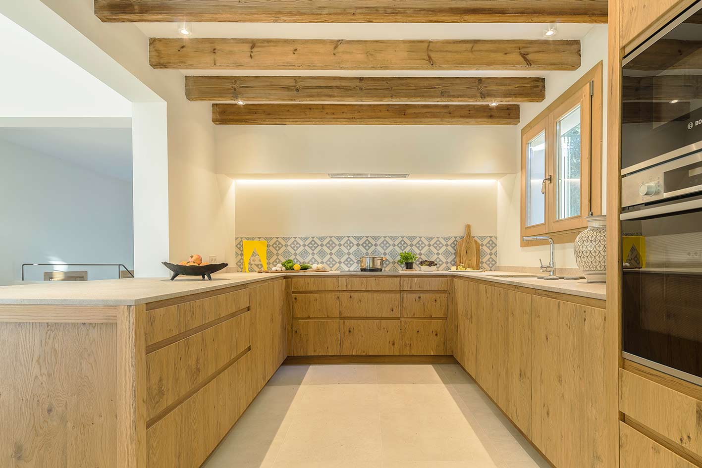 U-förmige Küche mit Holzschränken, heller Arbeitsplatte, sichtbaren Balken und gemustertem Fliesenspiegel