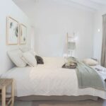 Weißes Schlafzimmer mit schräger Balkendecke, strukturierter Tagesdecke, grünem Plaid und Dekoleiter