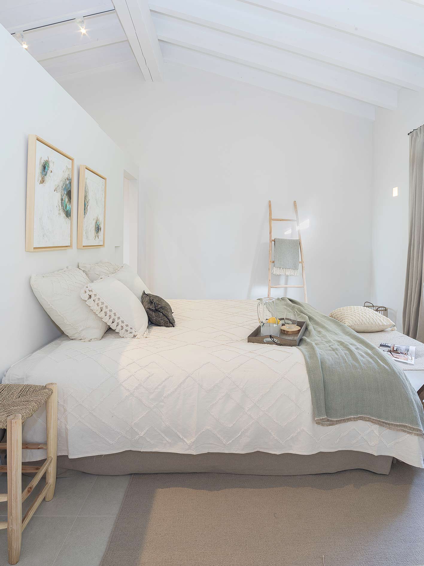 Weißes Schlafzimmer mit schräger Balkendecke, strukturierter Tagesdecke, grünem Plaid und Dekoleiter
