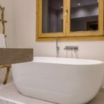 Detail einer weißen freistehenden Badewanne unter einem Holzfenster, mit Wandarmatur und Leiter aus Ästen