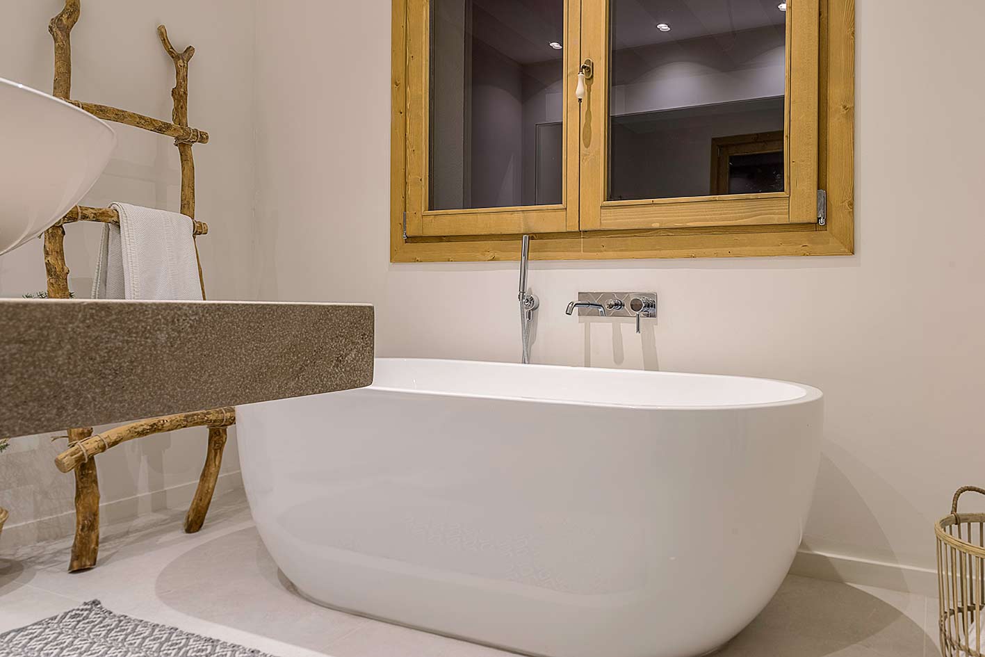 Detail einer weißen freistehenden Badewanne unter einem Holzfenster, mit Wandarmatur und Leiter aus Ästen
