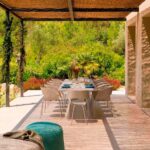 Essbereich im Freien mit langem Tisch und beigen Stühlen unter einer Schilfrohr-Pergola, türkisfarbener Pouf, Garten dahinter
