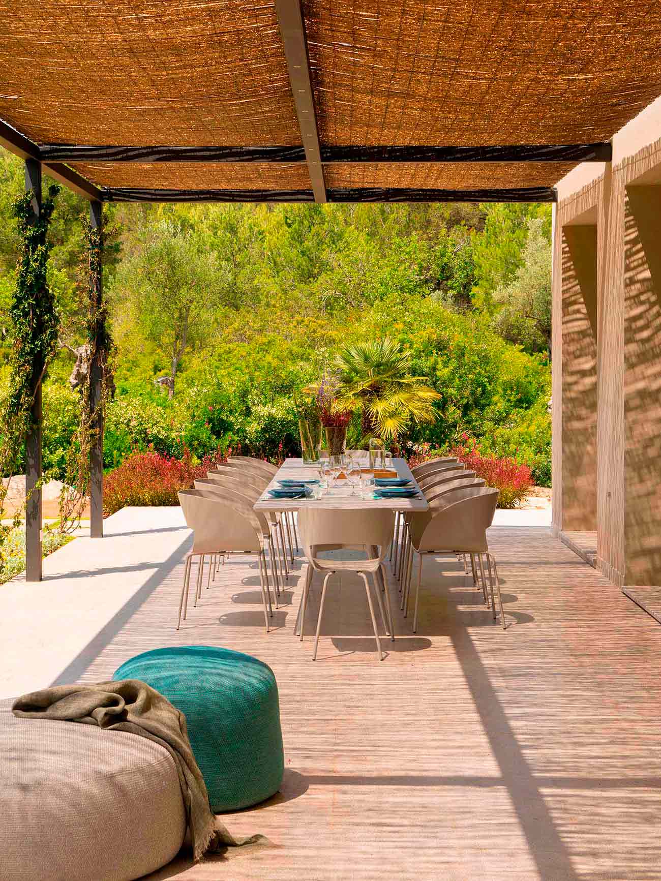 Essbereich im Freien mit langem Tisch und beigen Stühlen unter einer Schilfrohr-Pergola, türkisfarbener Pouf, Garten dahinter