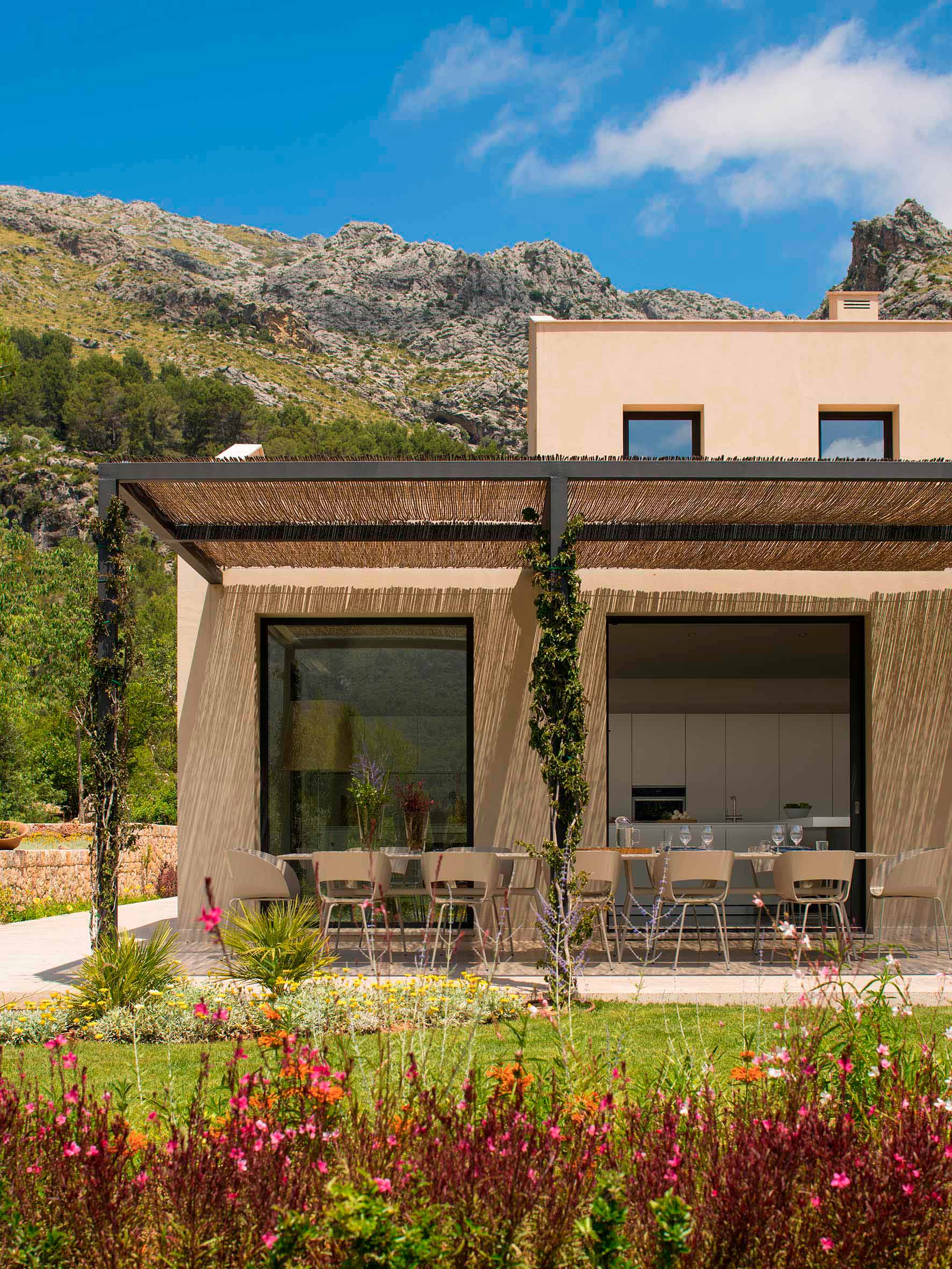 Fassade mit Schilfrohr-Pergola und großen Fenstern, blauer Himmel, Berg und rosa Blumen im Vordergrund