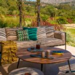 Detail des Outdoor-Sofas mit türkisfarbenen Kissen, senfgelber Decke und rundem Couchtisch aus Holz