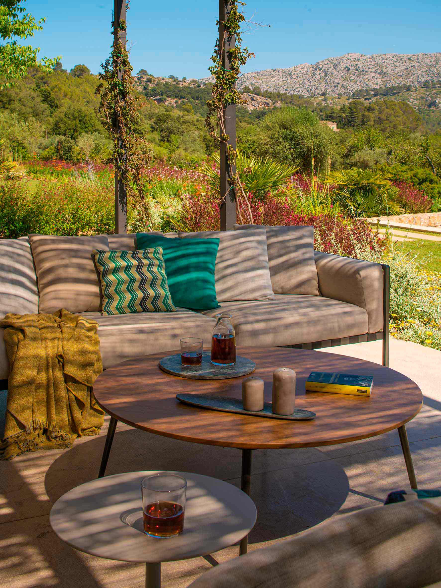 Detail des Outdoor-Sofas mit türkisfarbenen Kissen, senfgelber Decke und rundem Couchtisch aus Holz