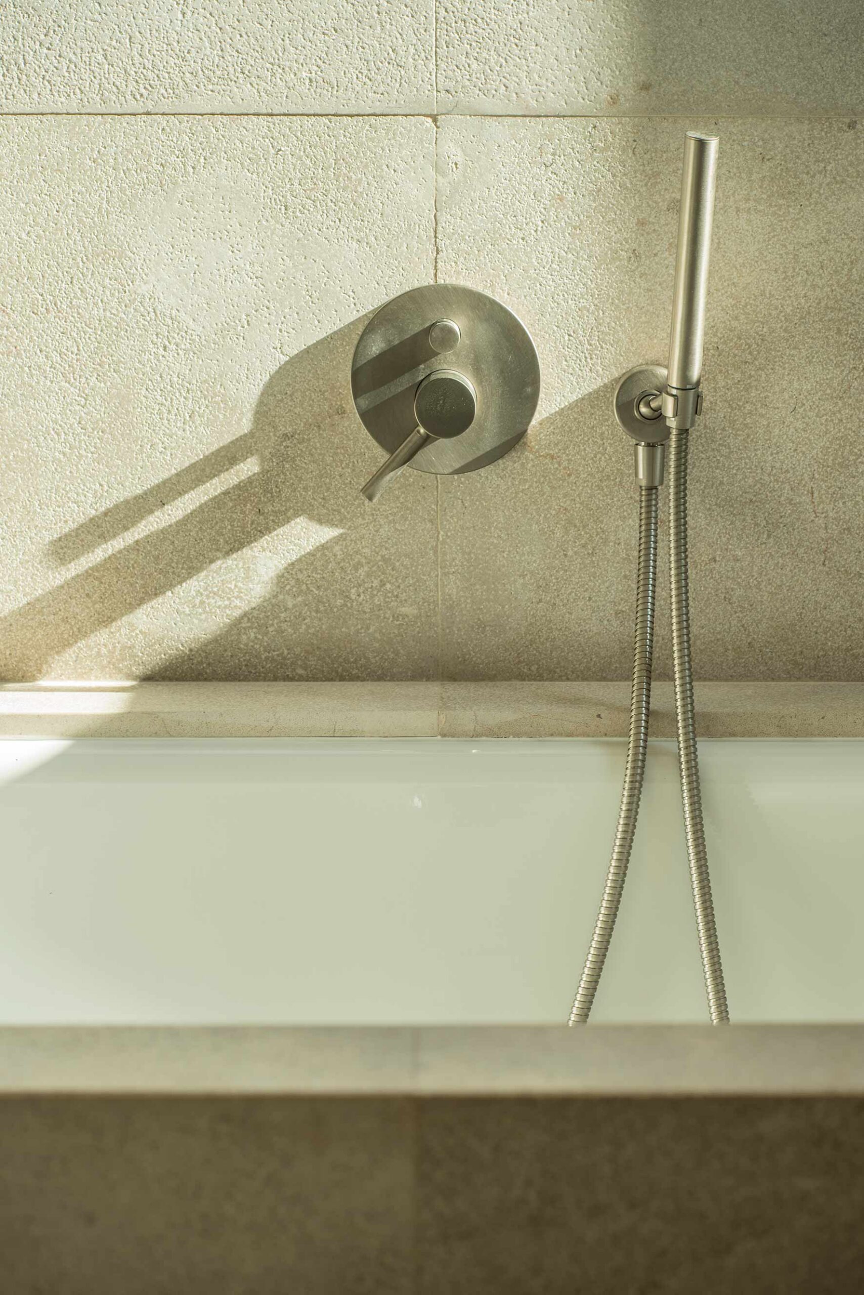 Detail der Wandarmatur und Handbrause über der Badewanne, mit heller Steinwand