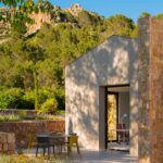 Verputzter Pavillon mit Steinmauer, Terrasse mit Tisch und gelben Stühlen und Hügel im Hintergrund