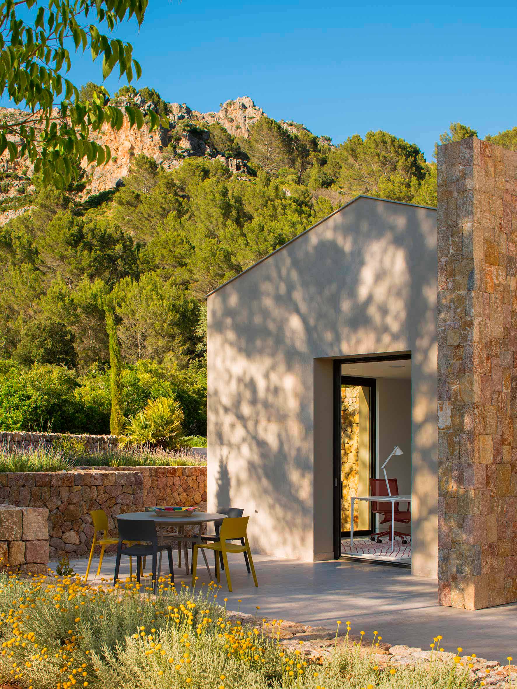 Verputzter Pavillon mit Steinmauer, Terrasse mit Tisch und gelben Stühlen und Hügel im Hintergrund
