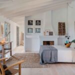 Bedroom with sloping white-beamed ceiling, fireplace, bed with green cushions, jute rug and pale parquet