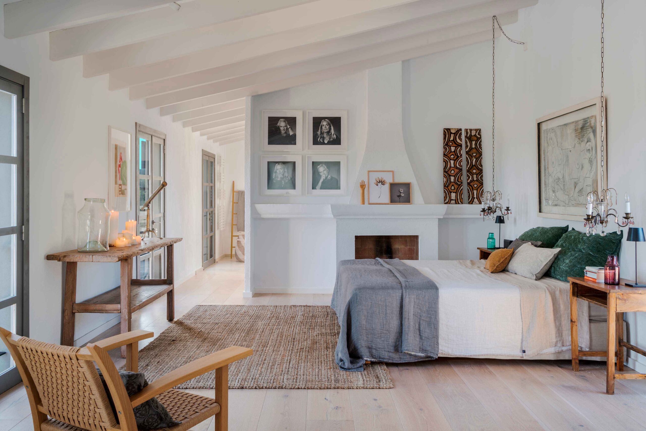 Bedroom with sloping white-beamed ceiling, fireplace, bed with green cushions, jute rug and pale parquet