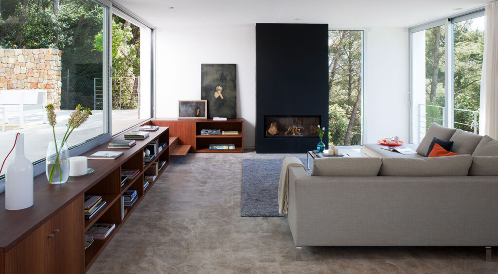 Salón con chimenea negra empotrada, estantería de madera, sofá gris y ventanales al bosque