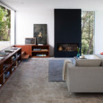 Salón con chimenea negra empotrada, estantería de madera, sofá gris y ventanales al bosque