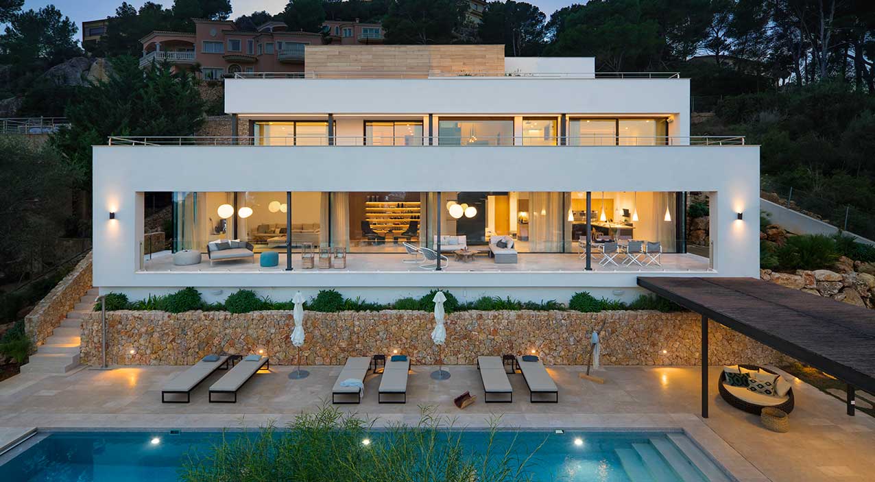 Fachada de Gotmar 142 al anochecer con muro de piedra, piscina y salón acristalado iluminado