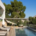 Piscina alargada con tumbonas, pérgola de sombra, barandilla de cristal y montaña al fondo