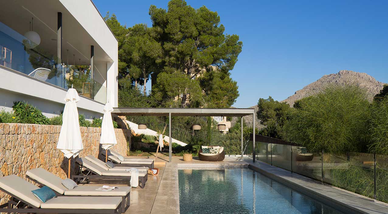 Piscina alargada con tumbonas, pérgola de sombra, barandilla de cristal y montaña al fondo