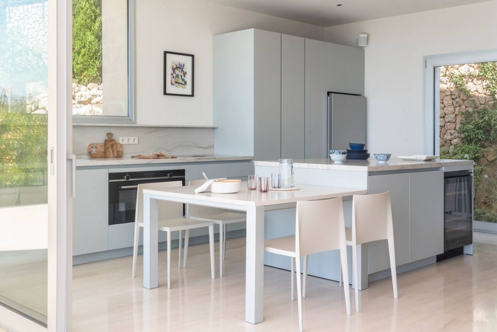 Cocina con armarios gris azulado, isla con encimera clara, mesa blanca con sillas y ventanal corredero