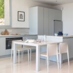 Cocina con armarios gris azulado, isla con encimera clara, mesa blanca con sillas y ventanal corredero