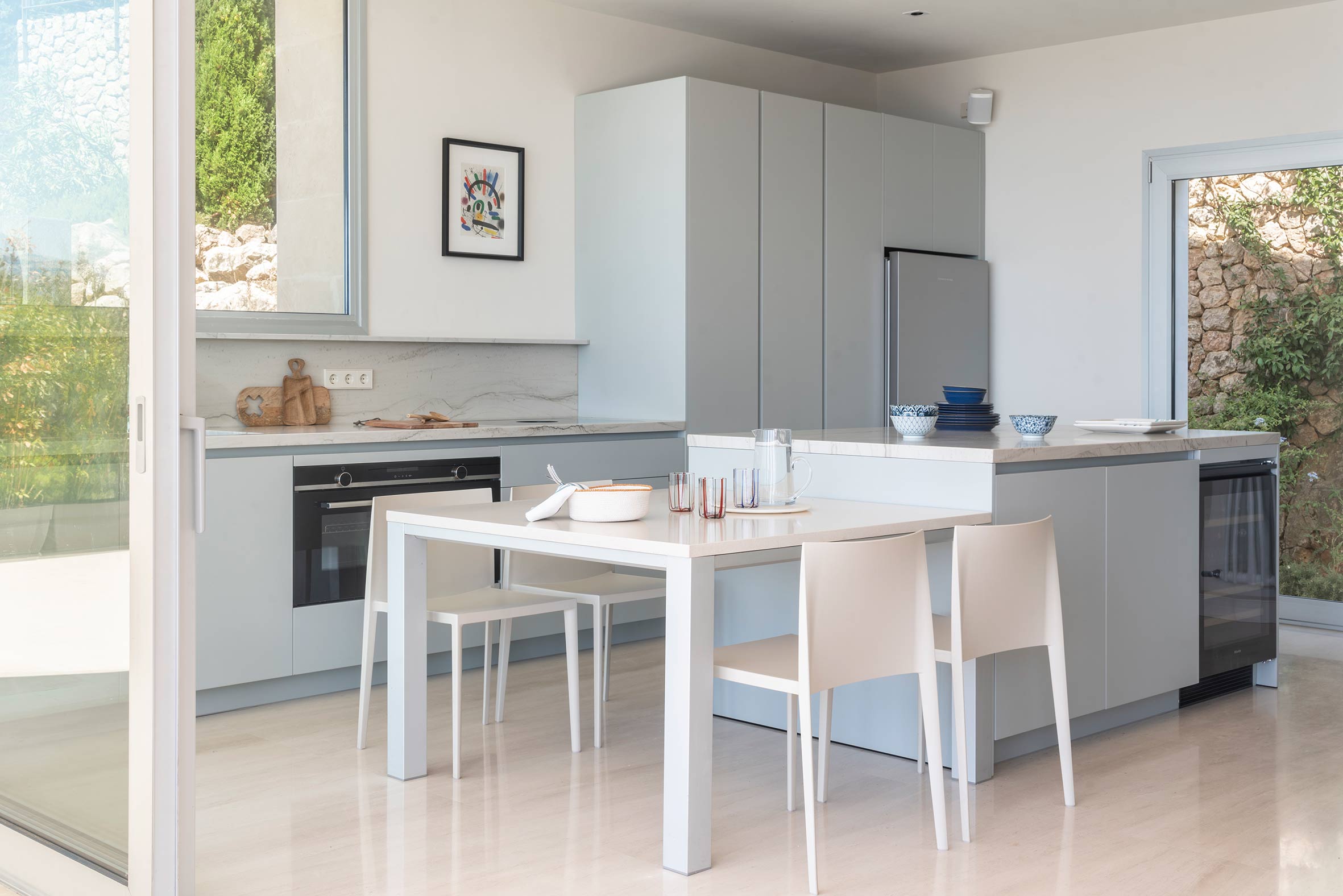Cocina con armarios gris azulado, isla con encimera clara, mesa blanca con sillas y ventanal corredero