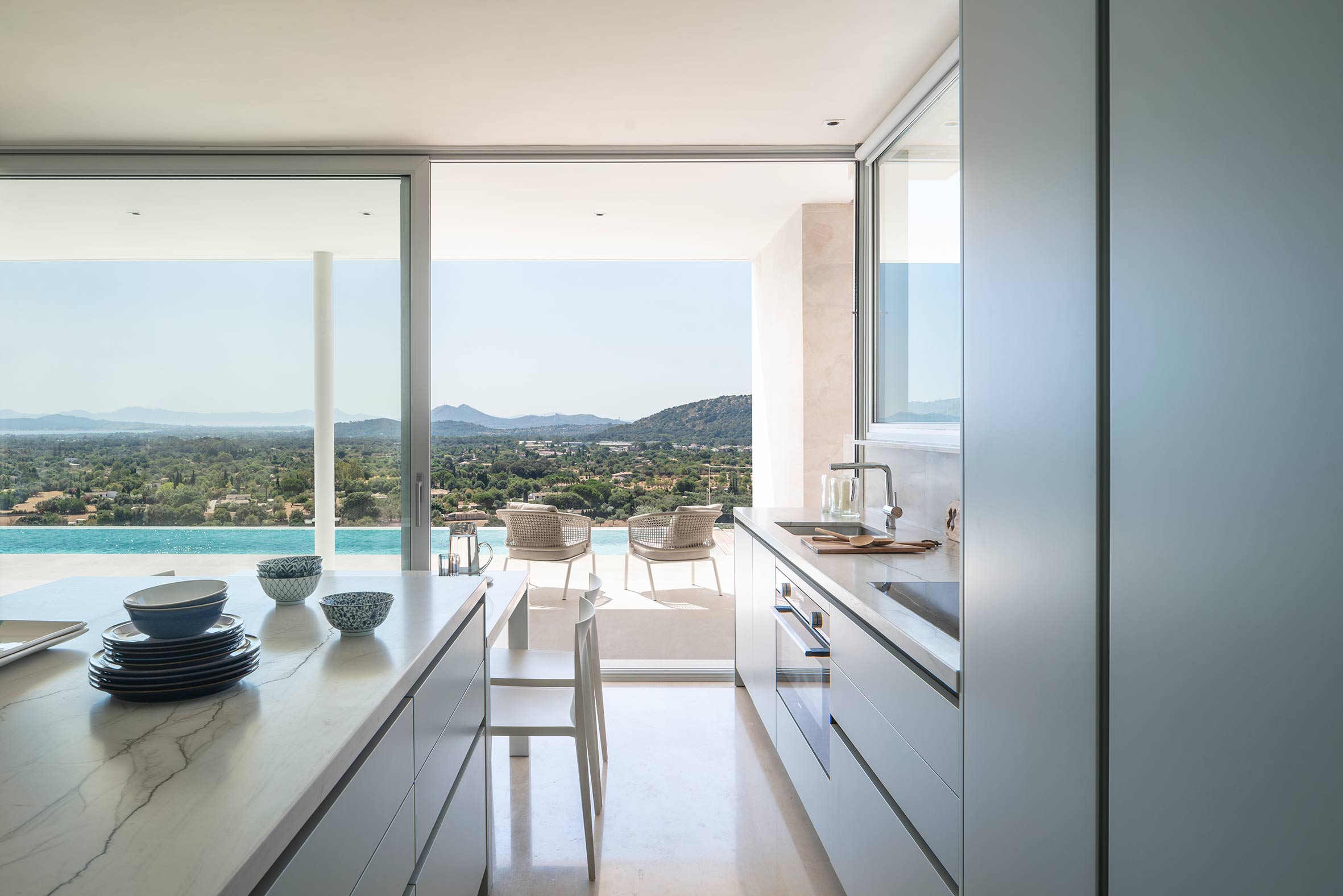 Cocina abierta a la terraza, isla con encimera veteada y vistas a la piscina y al valle