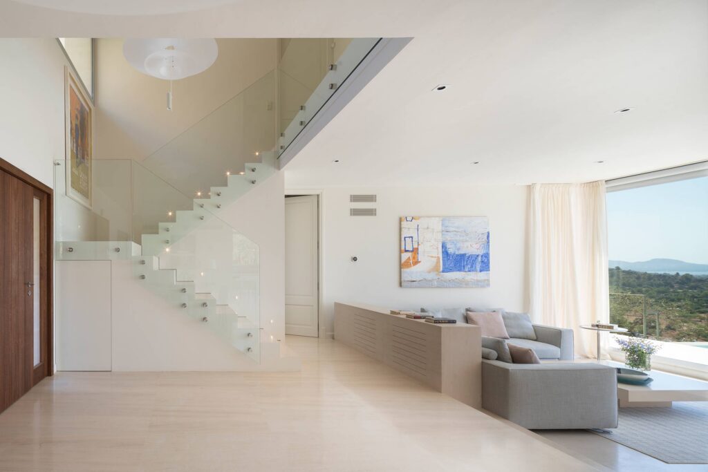 Salón luminoso con escalera acristalada, suelo de piedra clara, sofá gris y gran ventanal con vistas