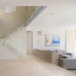 Salón luminoso con escalera acristalada, suelo de piedra clara, sofá gris y gran ventanal con vistas