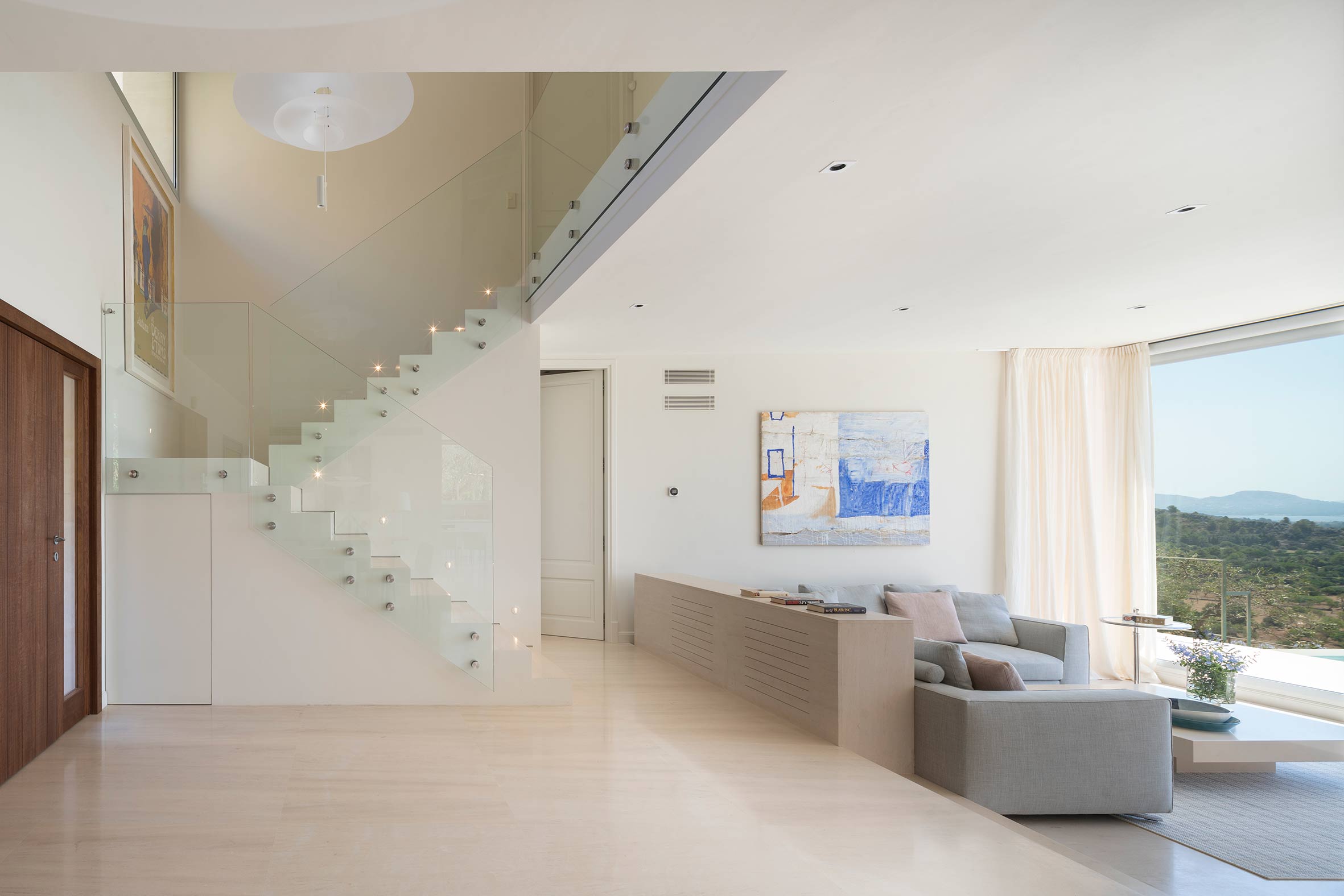 Salón luminoso con escalera acristalada, suelo de piedra clara, sofá gris y gran ventanal con vistas