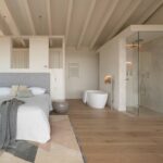 Dormitorio principal con cabecero gris, bañera exenta, ducha acristalada y suelo de madera bajo vigas claras