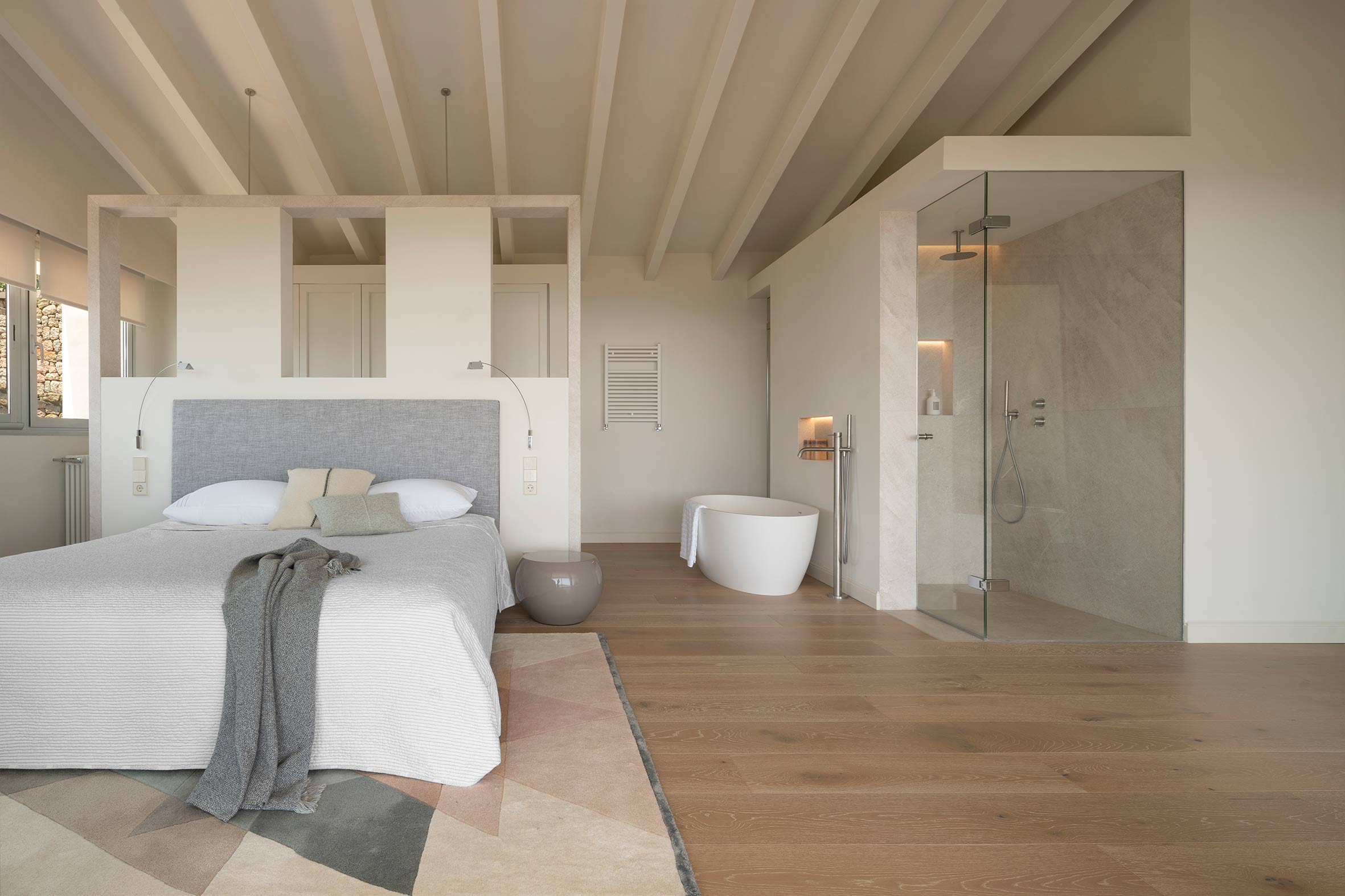 Dormitorio principal con cabecero gris, bañera exenta, ducha acristalada y suelo de madera bajo vigas claras
