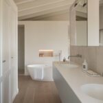 Baño con bañera exenta, doble lavabo sobre encimera blanca, revestimiento efecto piedra y suelo de madera